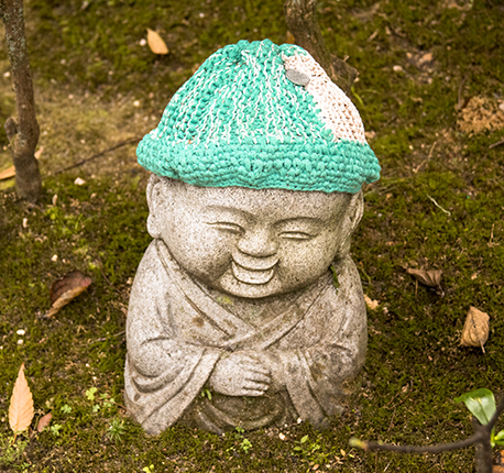 Pequeño Jizo de granito, sonriente, manos juntas, con gorro de  hilo color verde agua y blanco. 