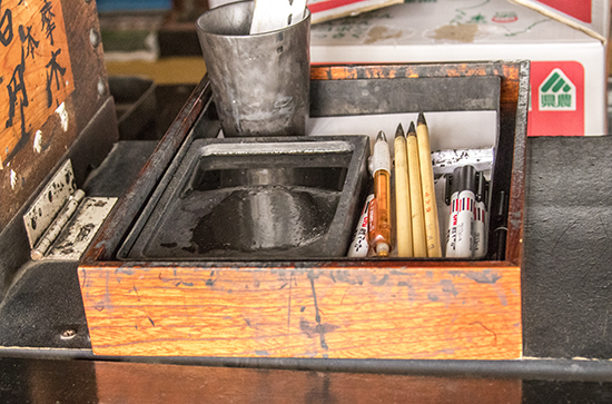 pinceles, tinta, un vaso, una caja d emadera abierta con kanji, un lápiz, algún rotulador negro